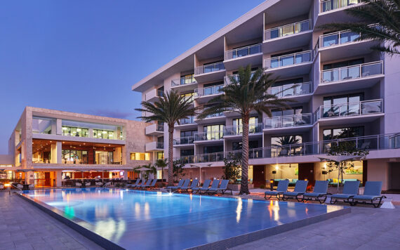 A modern hotel with multiple stories overlooks a serene swimming pool lined with lounge chairs. Tall palm trees add a tropical feel to the scene, set against a clear evening sky with warm lights glowing from the building.