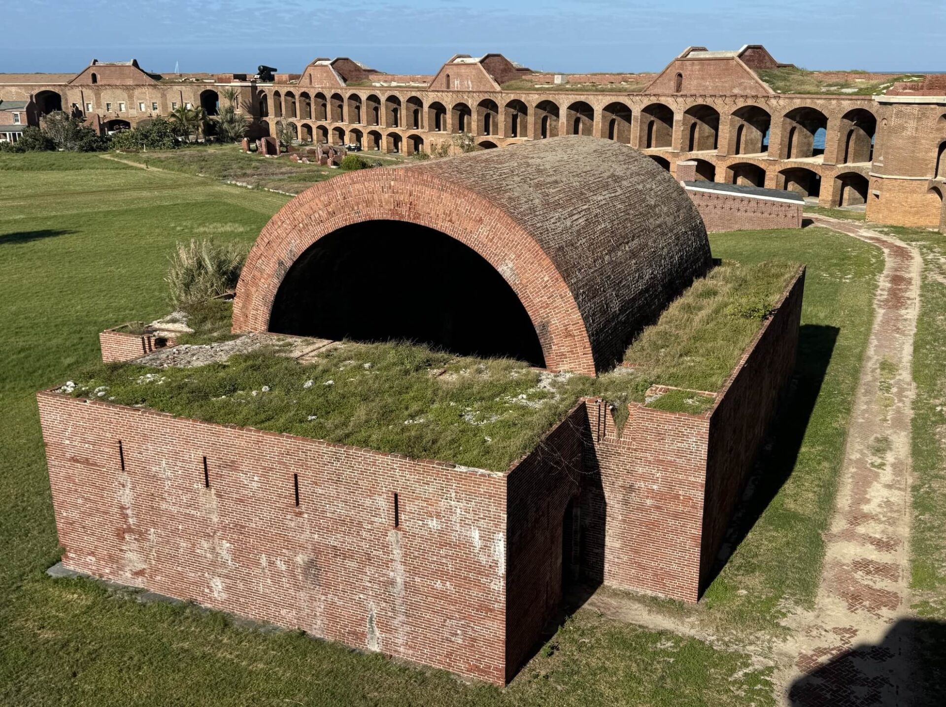 An old brick fort structure stands proudly under a large arched roof covered in grass at Dry Tortugas National Park. Surrounding it is grassy terrain, meandering paths, and other charming brick buildings with archways. The sky above is a serene blue dotted with a few clouds.