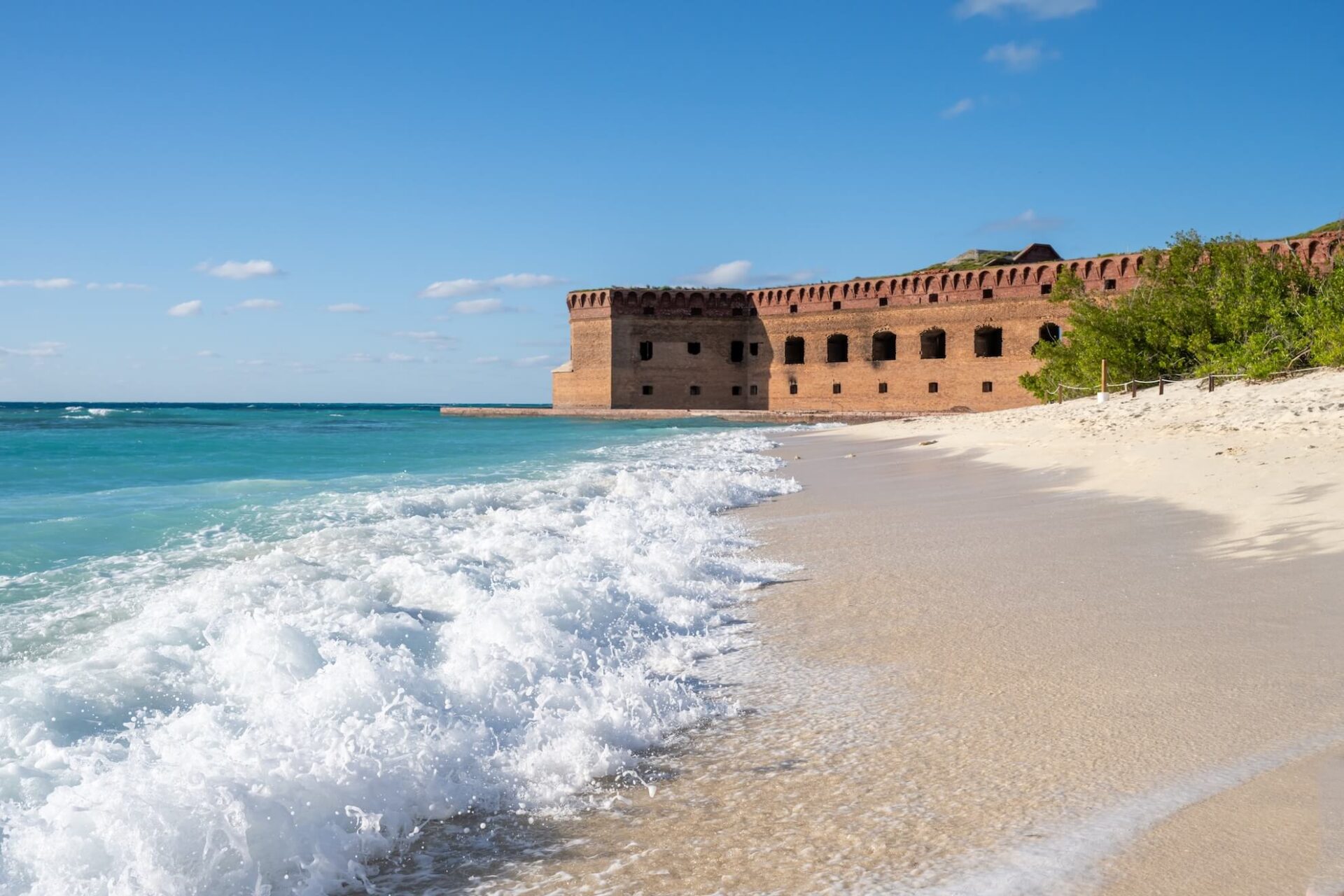 Sunny beach with gentle waves washing over the sand beside a large, historic brick fort at Dry Tortugas National Park. The blue sky is dotted with a few clouds, and sparse greenery grows near the forts walls.