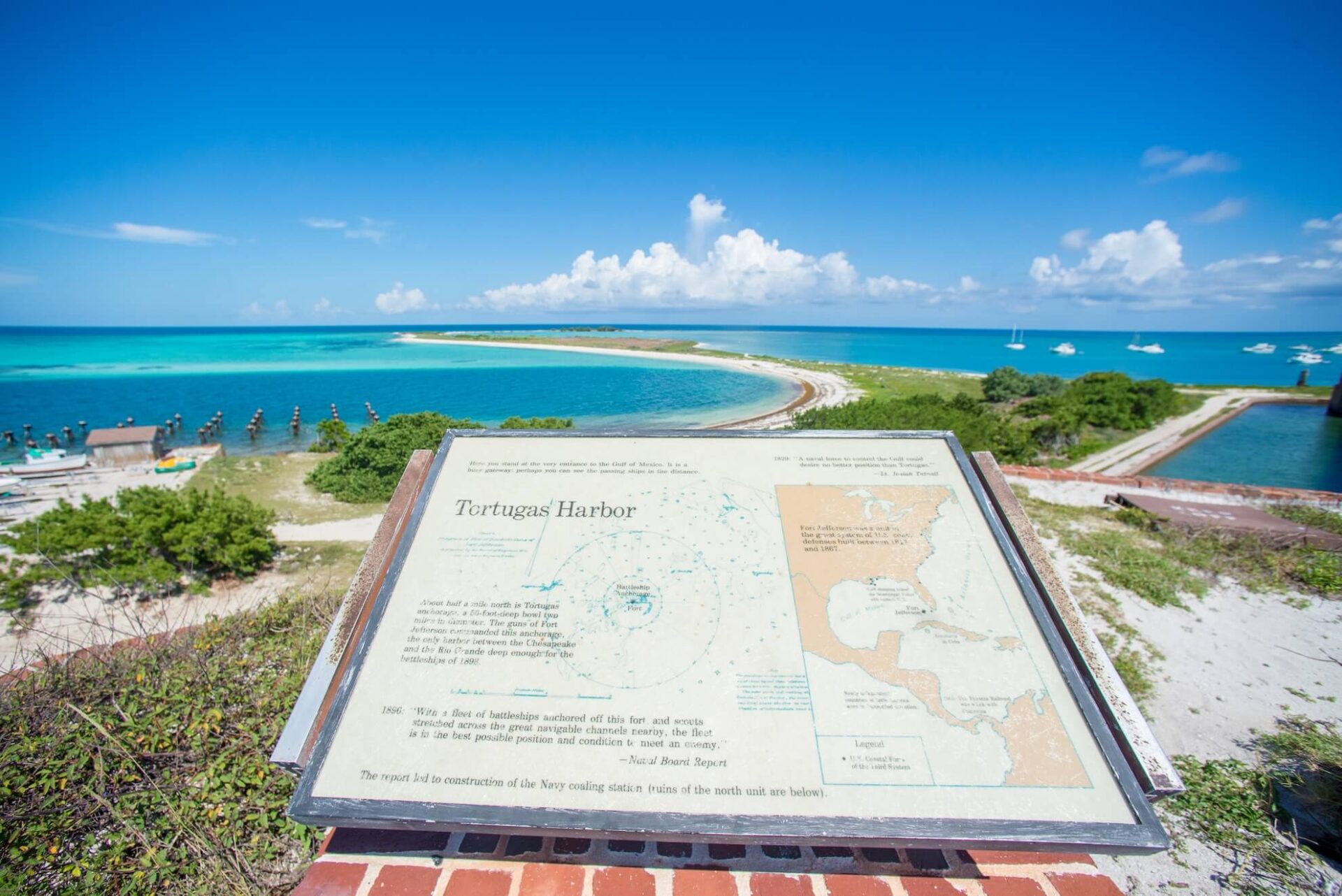 A scenic view of a coastal area features a clear blue sky, turquoise waters, and boats anchored near the shore. In the foreground, an informational plaque about Tortugas Harbor stands proudly amid green shrubs and sandy paths, inviting visitors to explore Dry Tortugas National Park.