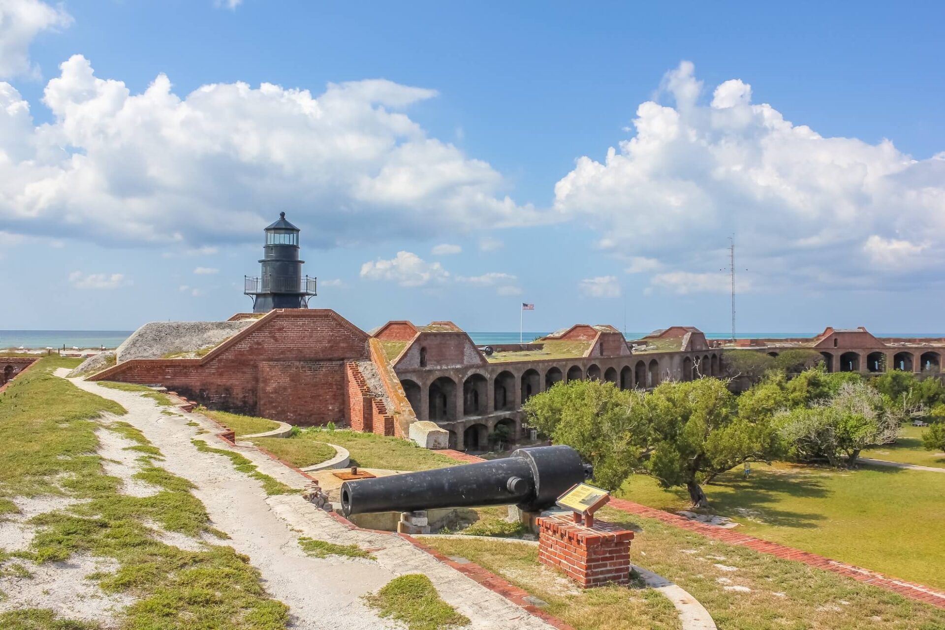 Historic fort with brick structures and arches near the ocean in Dry Tortugas National Park. A black lighthouse rises above the fort, while a large cannon is prominently displayed on the grassy area. The sky is blue with scattered clouds, adding to this picturesque scene.