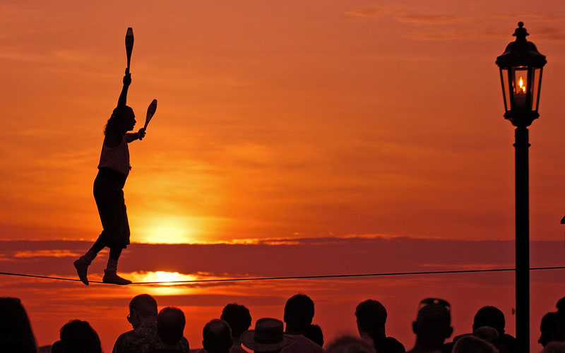 Silhouette of a person juggling while walking on a tightrope at sunset, ticking off a daring feat from their summer bucket list. A crowd watches below, and a street lamp is visible on the right. The sky is a vibrant orange with scattered clouds.