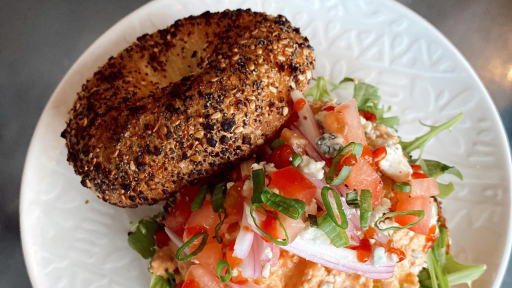 A toasted everything bagel sits on a plate beside a fresh salad of arugula, diced tomatoes, red onions, green onions, and crumbled cheese—a delicious pause between your favorite things to do in Lake George.