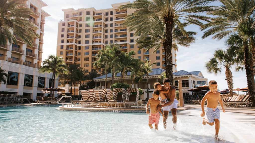 A man and two children play in a sunny resort pool with palm trees and a tall hotel building in the background. All three are smiling and wearing swimwear.