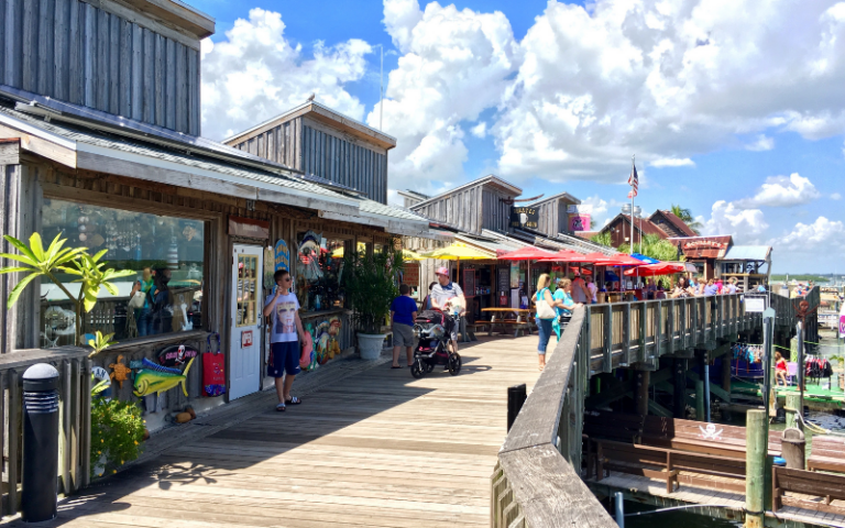 People stroll along a wooden boardwalk lined with shops, outdoor seating, and colorful umbrellas on a sunny day. The boardwalk overlooks water, with clouds dotting the blue sky.