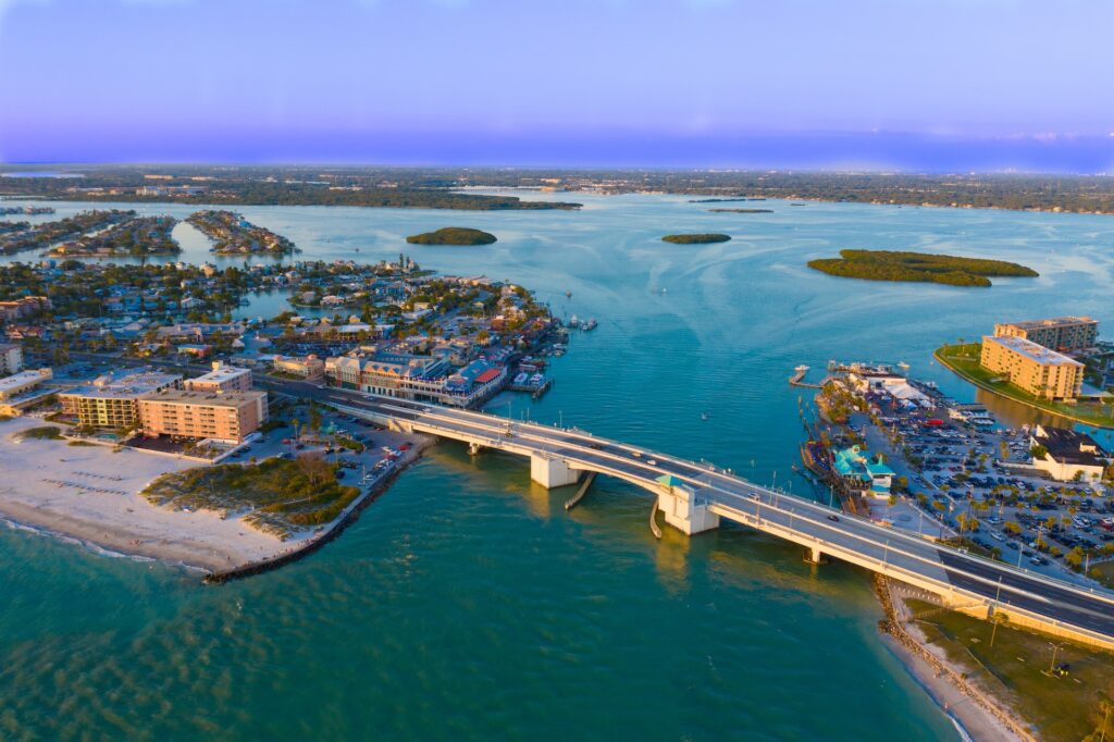 Aerial view of a coastal city with a bridge over turquoise water, small islands, sandy beaches, and waterfront buildings, taken during sunset.