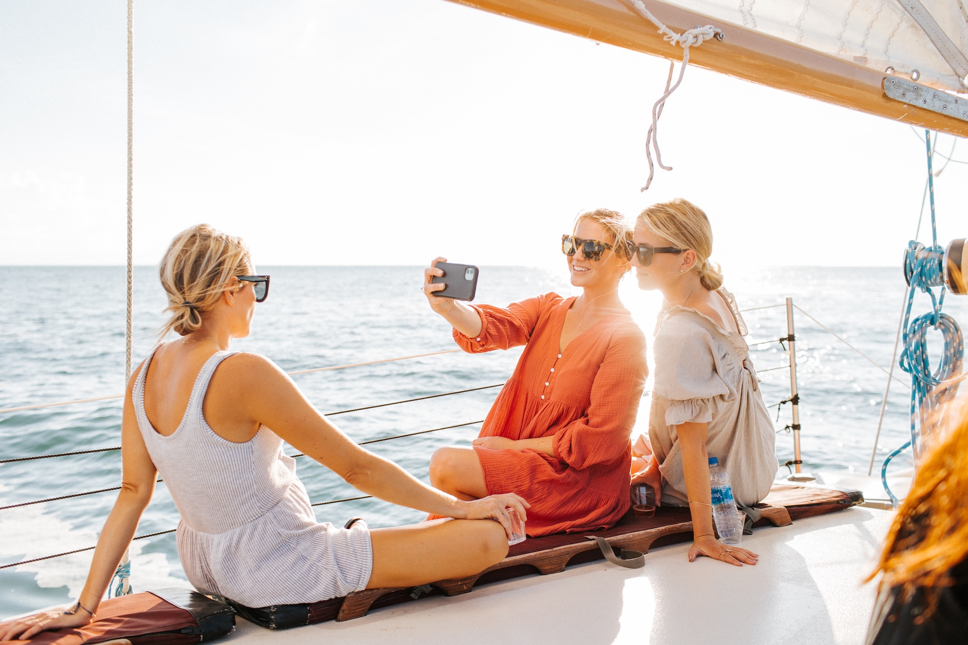 Three women in sunglasses sit on a sailboat, smiling and posing for a selfie together on a sunny day with the ocean in the background.