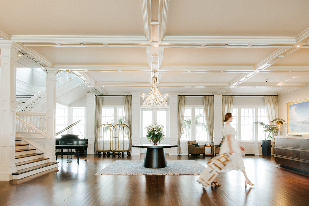 A woman in a white dress walks through a bright hotel lobby with a piece of rolling luggage.