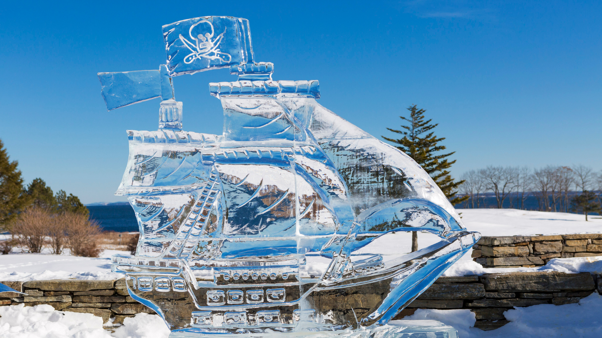 An intricate ice sculpture of a pirate ship, complete with sails and a Jolly Roger flag, set outdoors on a snowy day. The background features a stone wall, trees, and a clear blue sky.