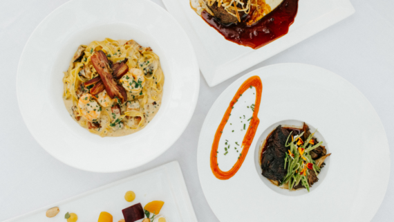 Overhead view of four elegantly plated gourmet dishes on white plates, including pasta with shrimp and herbs, beef with vegetables, a steak with sauce, and a dish with colorful garnishes, all on a white tablecloth.
