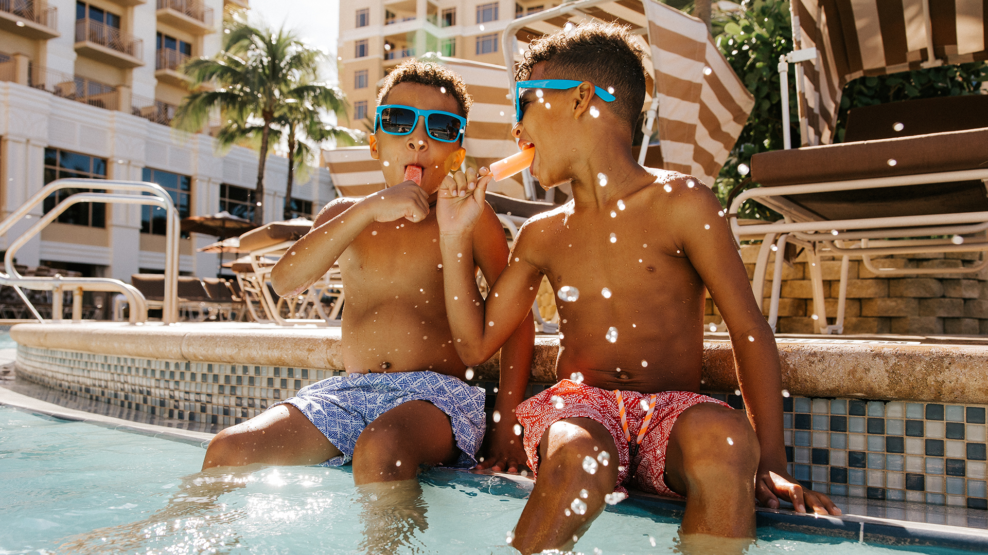 Two young boys wearing swim trunks and sunglasses sit by a pool, laughing and eating popsicles. Water droplets splash in front of them. Palm trees and lounge chairs are visible in the background.