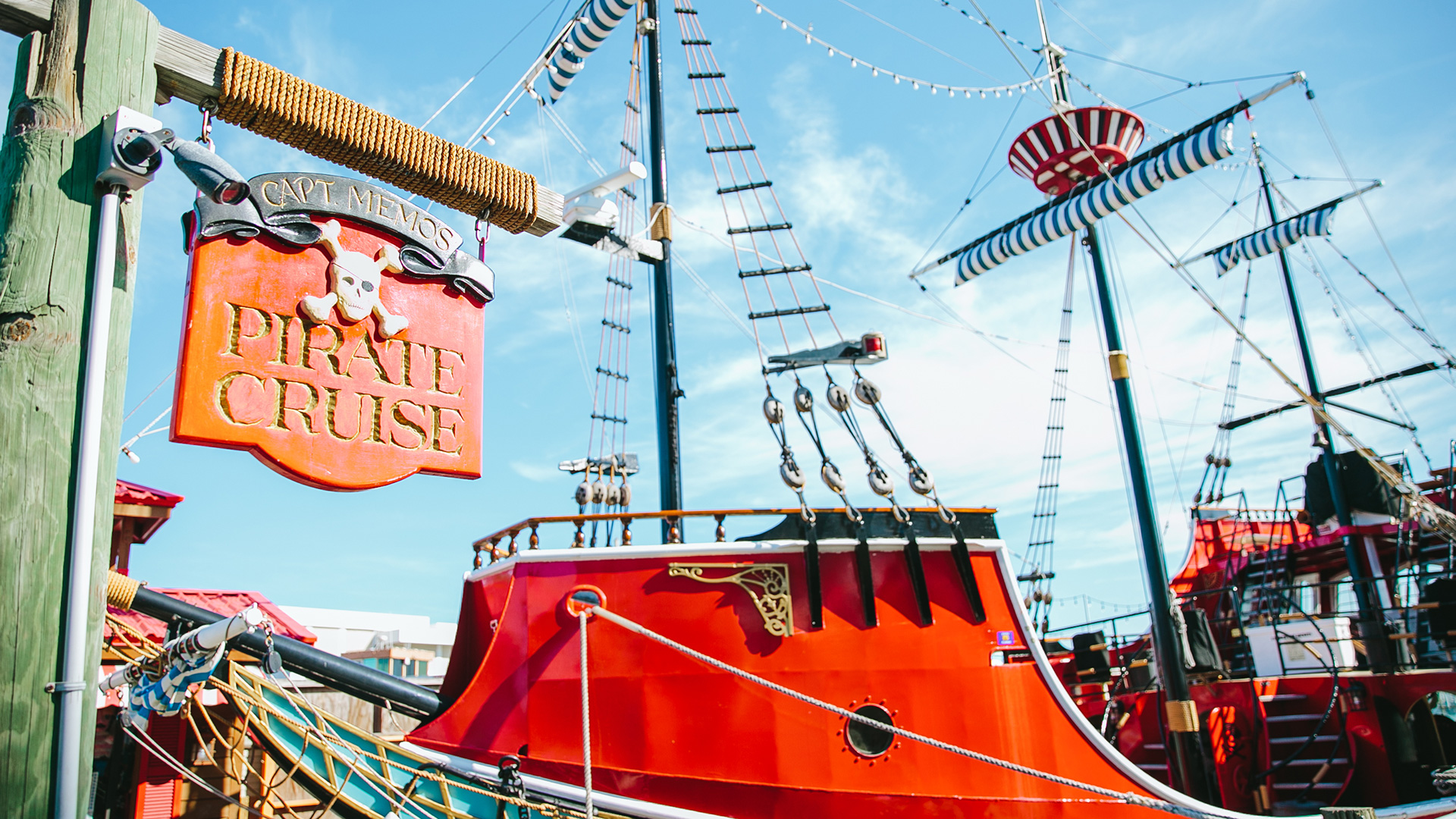 A bright red pirate-themed cruise ship docked, with a wooden sign reading PIRATE CRUISE featuring a skull and crossed swords. The ship has striped sails and decorative rigging against a blue sky.
