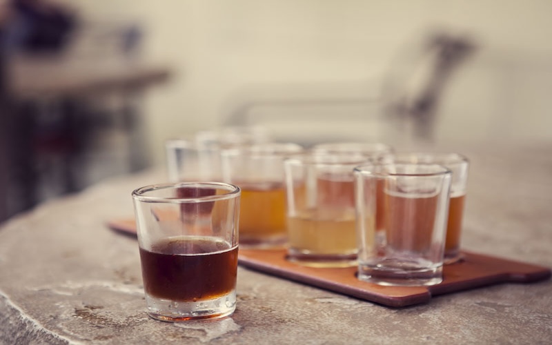 Several small glasses of beer or cider, each containing different shades of liquid, are arranged on a wooden tray atop a rustic, stone table. One glass stands slightly apart from the others in the foreground.