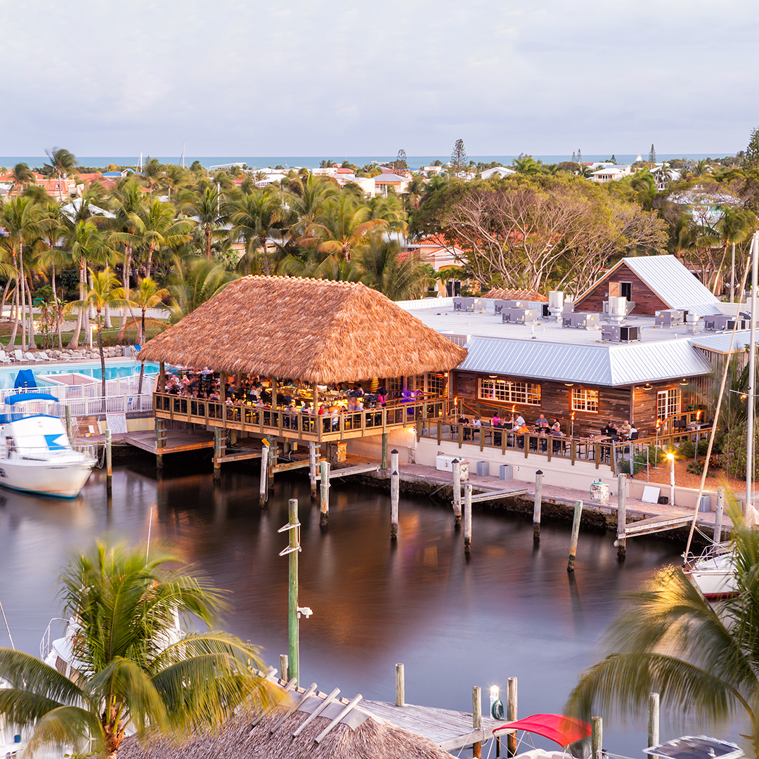 A waterfront restaurant with a thatched roof sits on a dock surrounded by palm trees, boats, and calm water, with houses and a distant view of the ocean in the background under a partly cloudy sky.