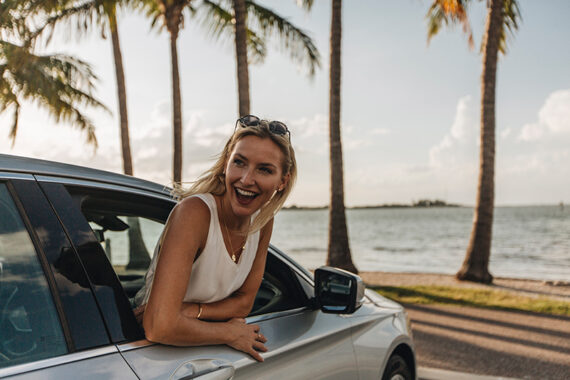 A smiling woman leans out of the window of a silver car parked by the beach, with palm trees and the ocean in the background on a sunny day.