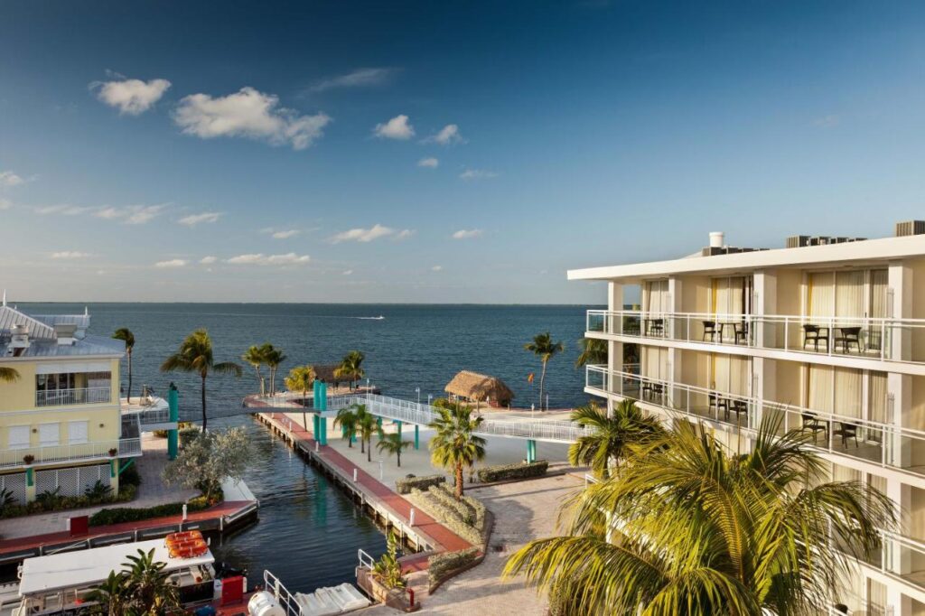 A scenic view of a coastal resort featuring modern buildings with balconies, a dock lined with palm trees, and a thatched-roof structure near the water. The ocean stretches out under a clear blue sky with a few clouds.