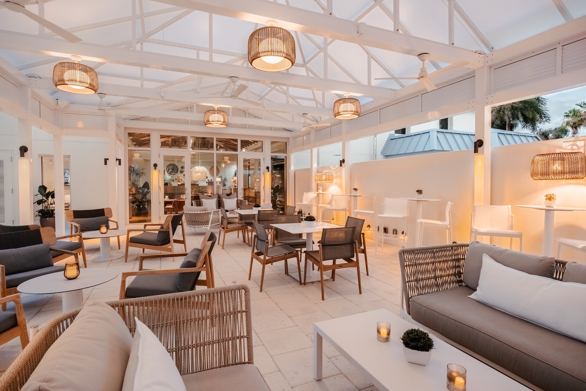 A stylish, modern outdoor patio with white beams, neutral-toned sofas and chairs, small tables, warm hanging lights, and candles, creating a cozy, inviting atmosphere for dining or relaxing.