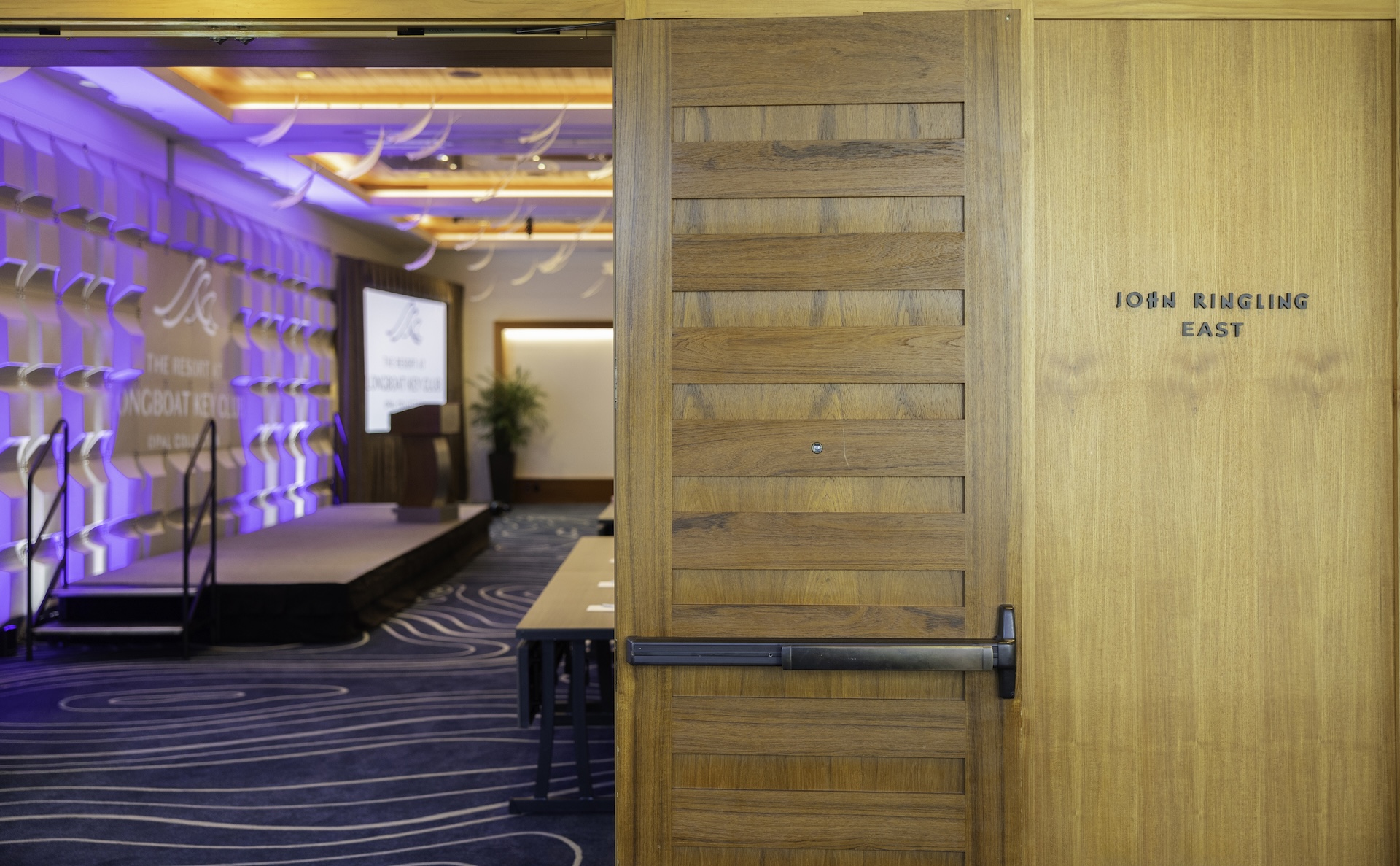 Open wooden door labeled John Ringling East reveals a modern conference room with a stage, tables, a projector screen, and blue accent lighting on the walls.