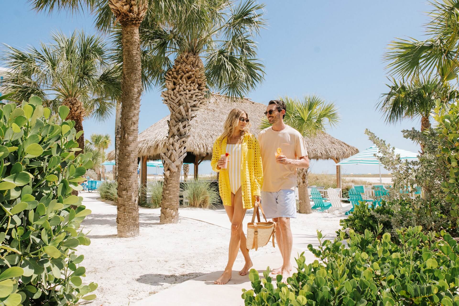 A couple walks barefoot on a sandy path lined with palm trees, wearing summer clothes and sunglasses, carrying drinks and a basket, with tiki huts and turquoise chairs in the background under a clear blue sky.