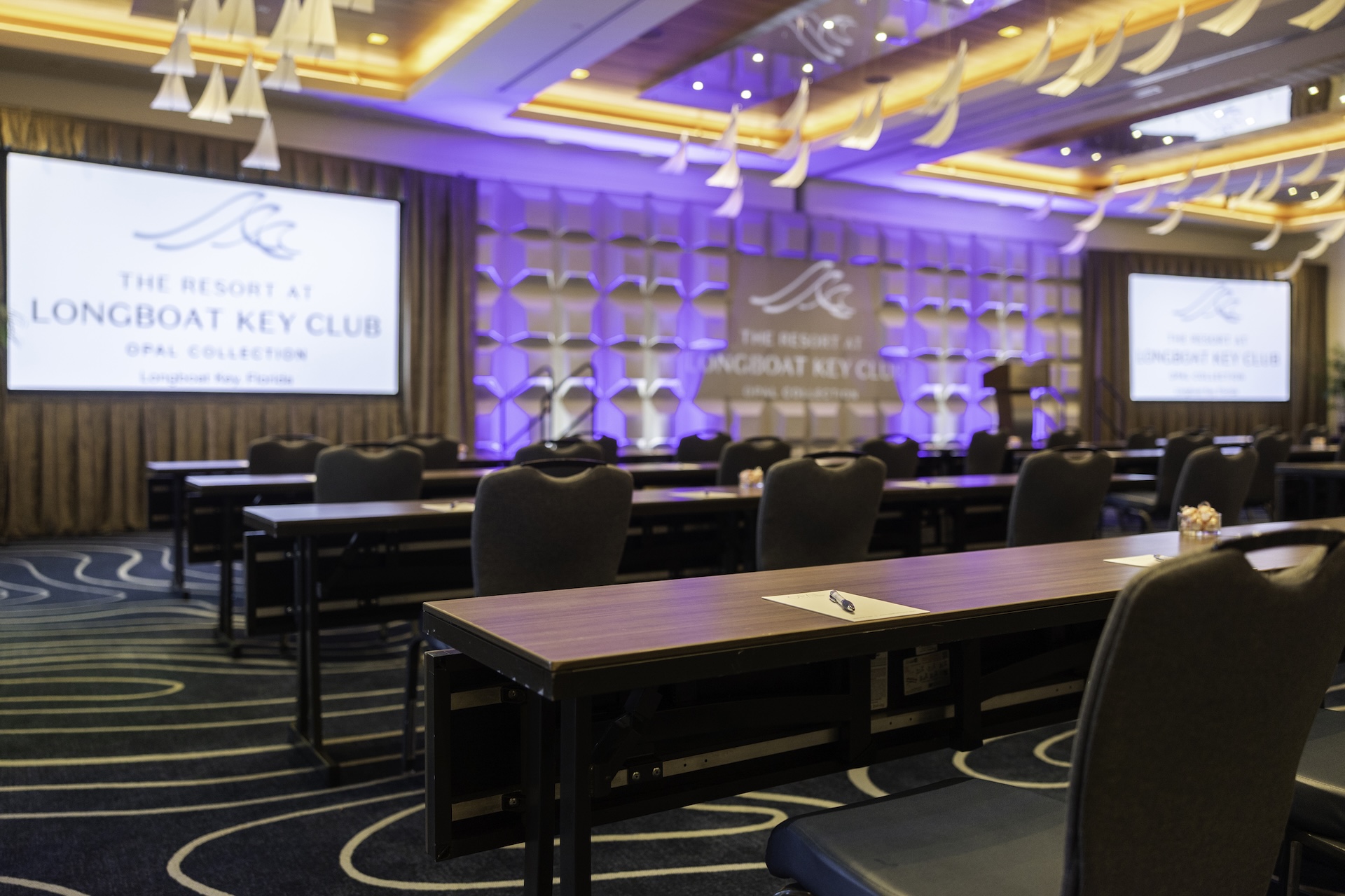 A conference room at The Resort at Longboat Key Club, set up with rows of tables and chairs facing large projection screens and a stage, under modern ceiling lights and decorations.