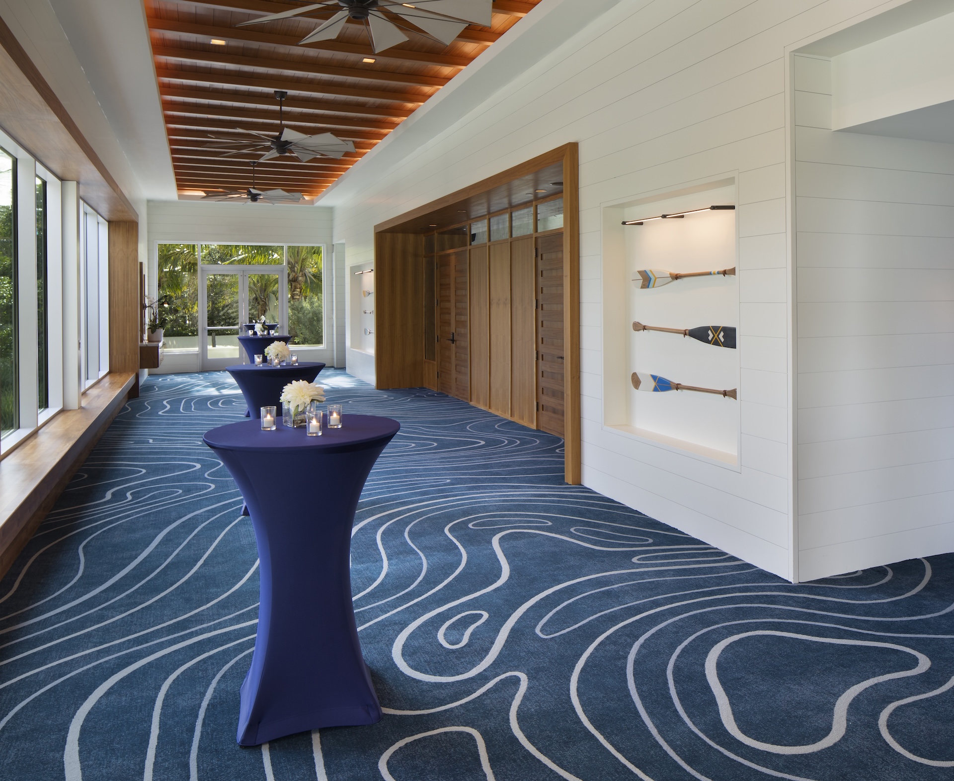A modern event space with blue patterned carpet, high wooden ceiling with fans, large windows, tall tables draped in navy cloth, and a wall display of decorative oars.