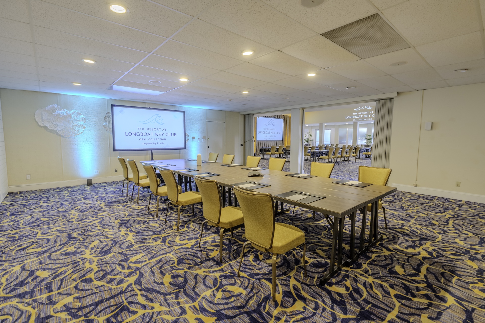 A conference room with yellow chairs arranged around rectangular tables, facing a screen displaying The Resort at Longboat Key Club. Blue accent lighting and patterned carpet decorate the space.