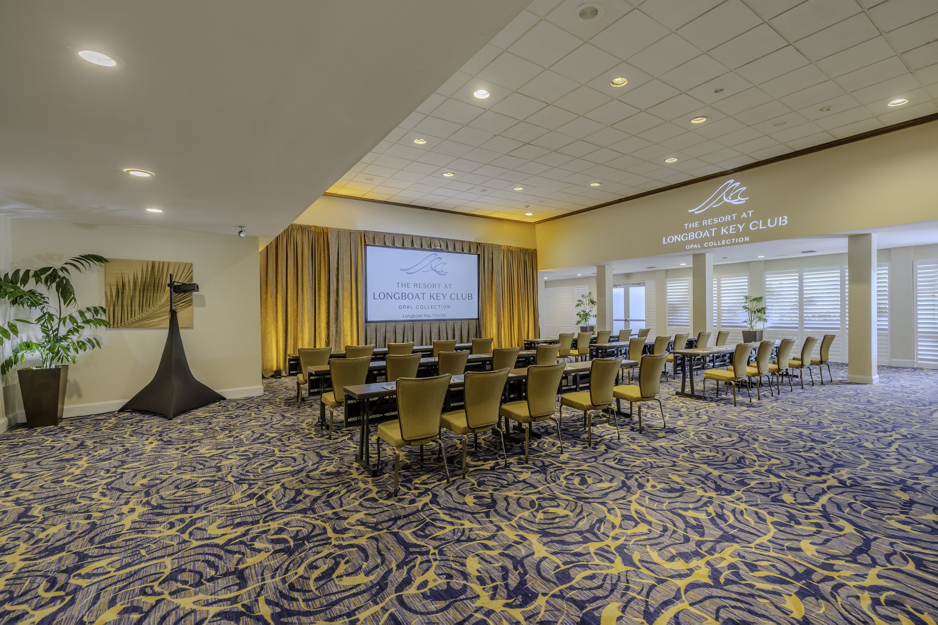 A conference room with rows of yellow chairs facing a projector screen and podium. The carpet has a blue and yellow pattern, and large windows let in natural light. The wall displays The Resort at Longboat Key Club.