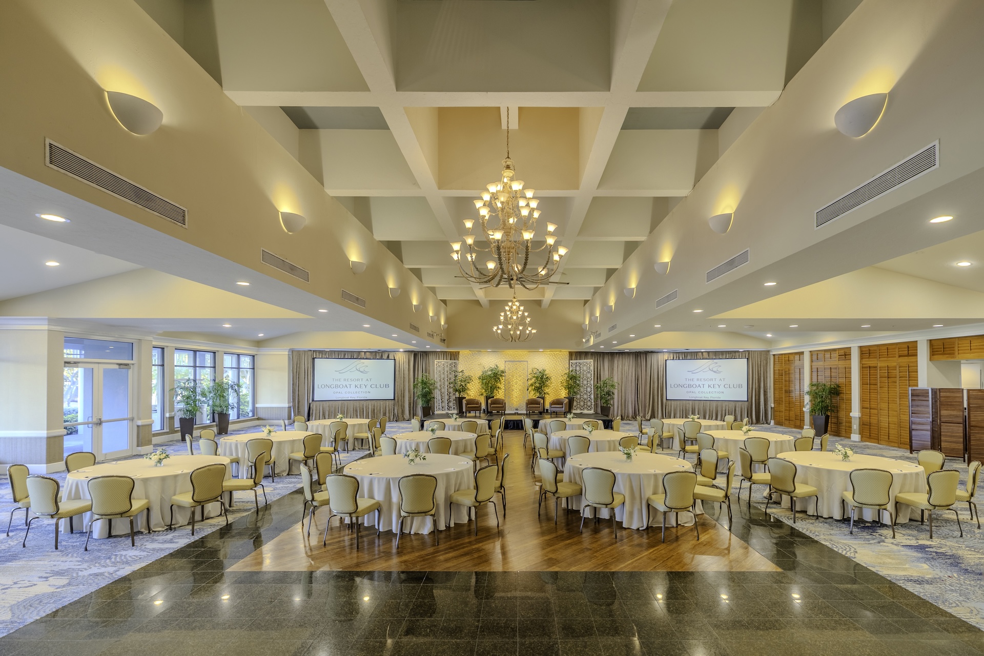 A spacious, elegant banquet hall with round tables and chairs arranged for an event, chandeliers hanging from a coffered ceiling, and large windows letting in natural light. Two large screens are visible at the far end.