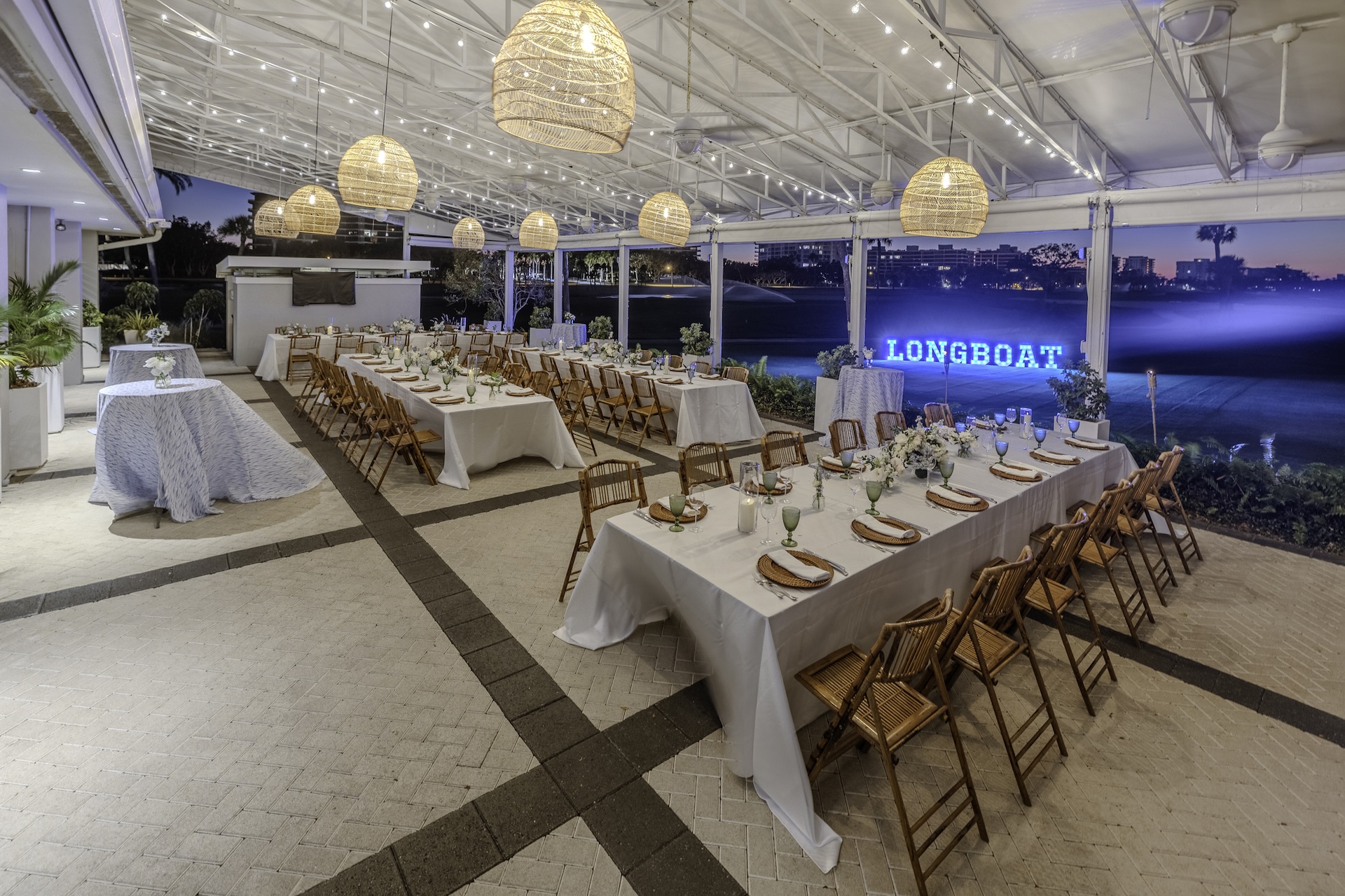 Outdoor event space with long tables set for dinner, wicker chairs, hanging woven lights, and a neon LONGBOAT sign in the background. The area is covered and overlooks a lawn at dusk.
