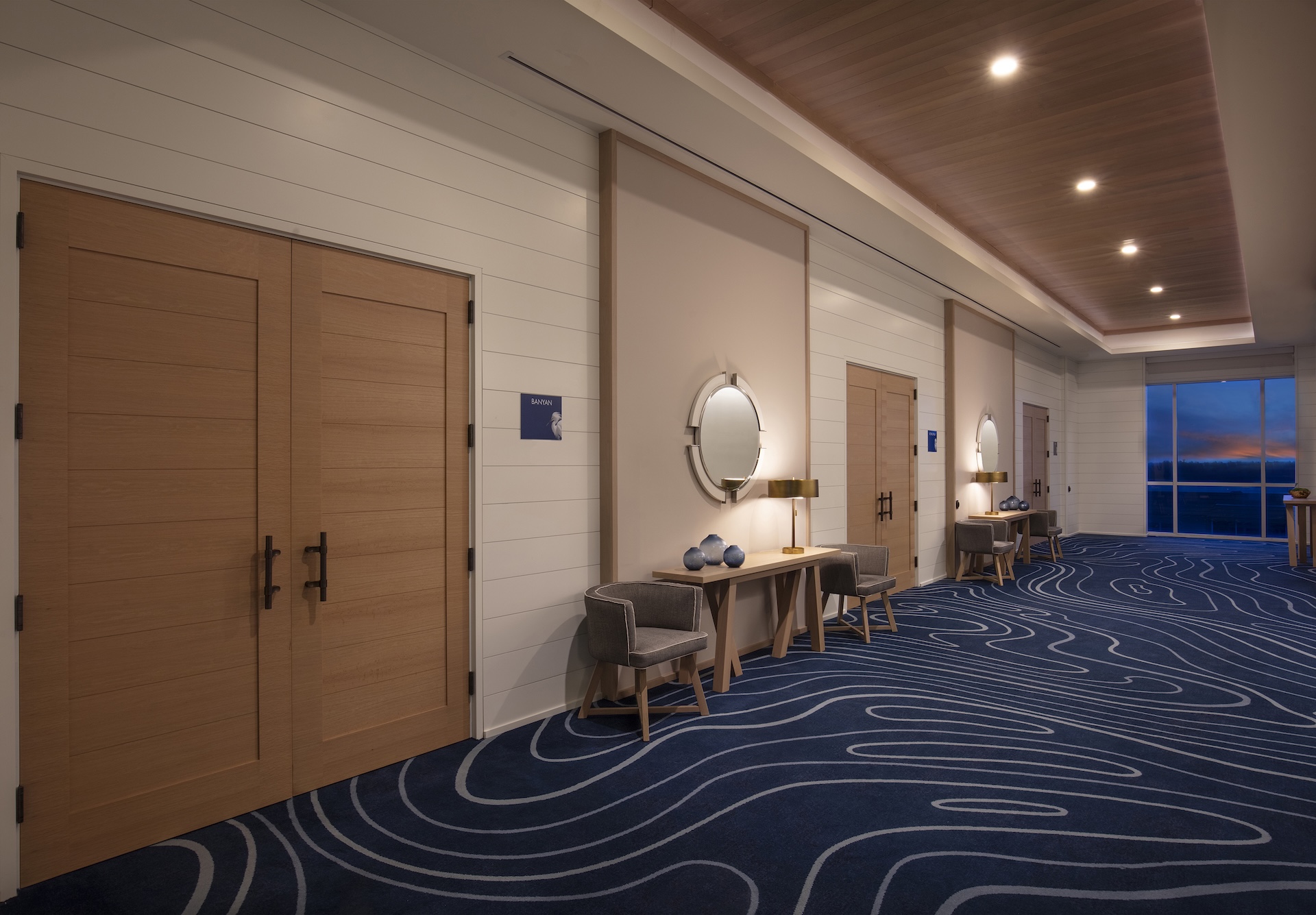 A modern hallway with blue carpet featuring wavy patterns, light wood double doors on the left, and console tables with round mirrors and decor along the wall. Large windows at the end show a sunset.