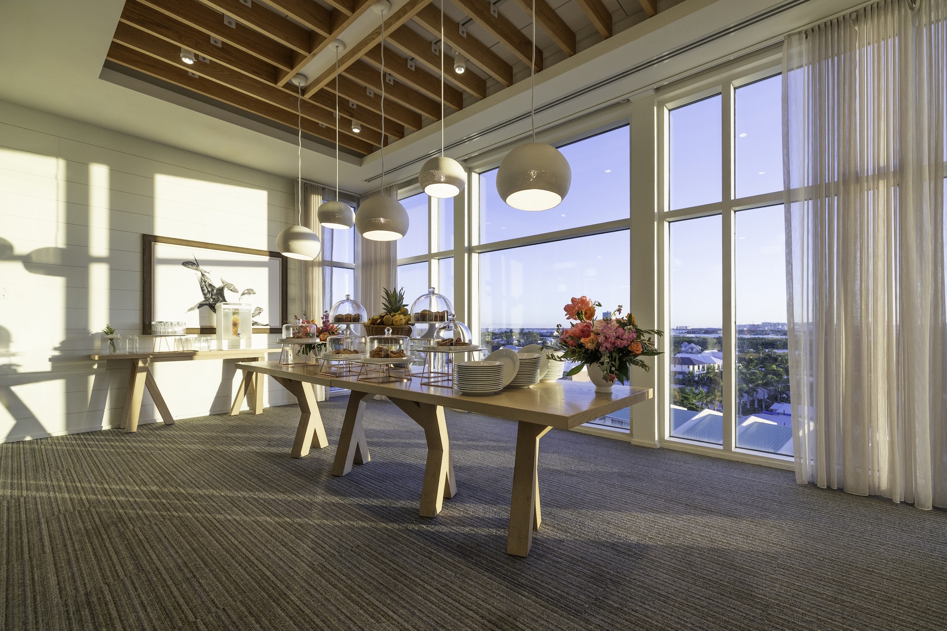 A bright, modern room with large windows, wooden ceiling beams, and pendant lights. Two wooden tables display plates, pastries, and floral arrangements, with sunlight streaming in and a scenic view outside.