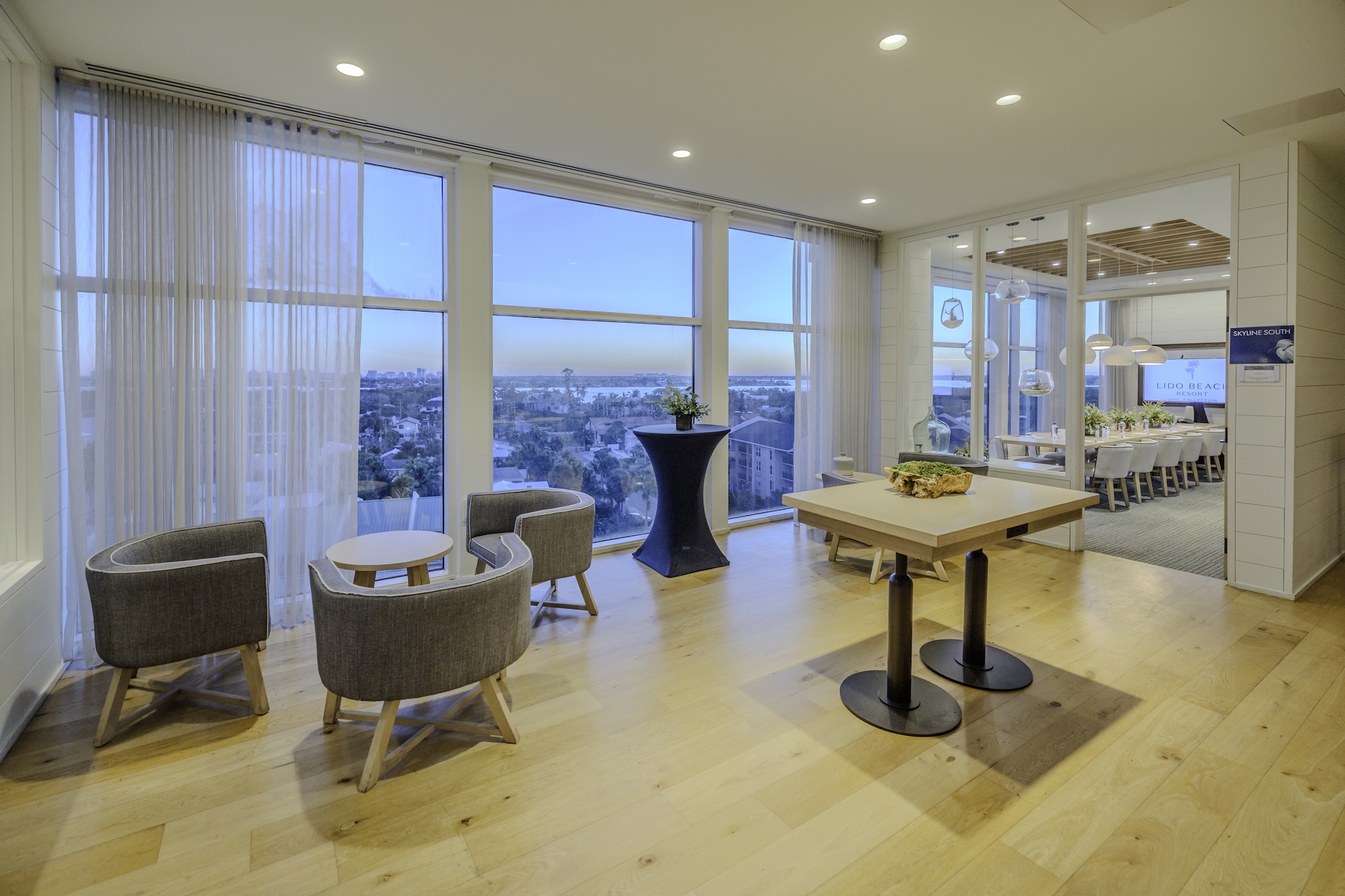 Modern lounge with light wood floors, gray chairs around small tables, and large windows offering a city view; a table with food and drinks is in the center, and a meeting room is visible through glass walls.