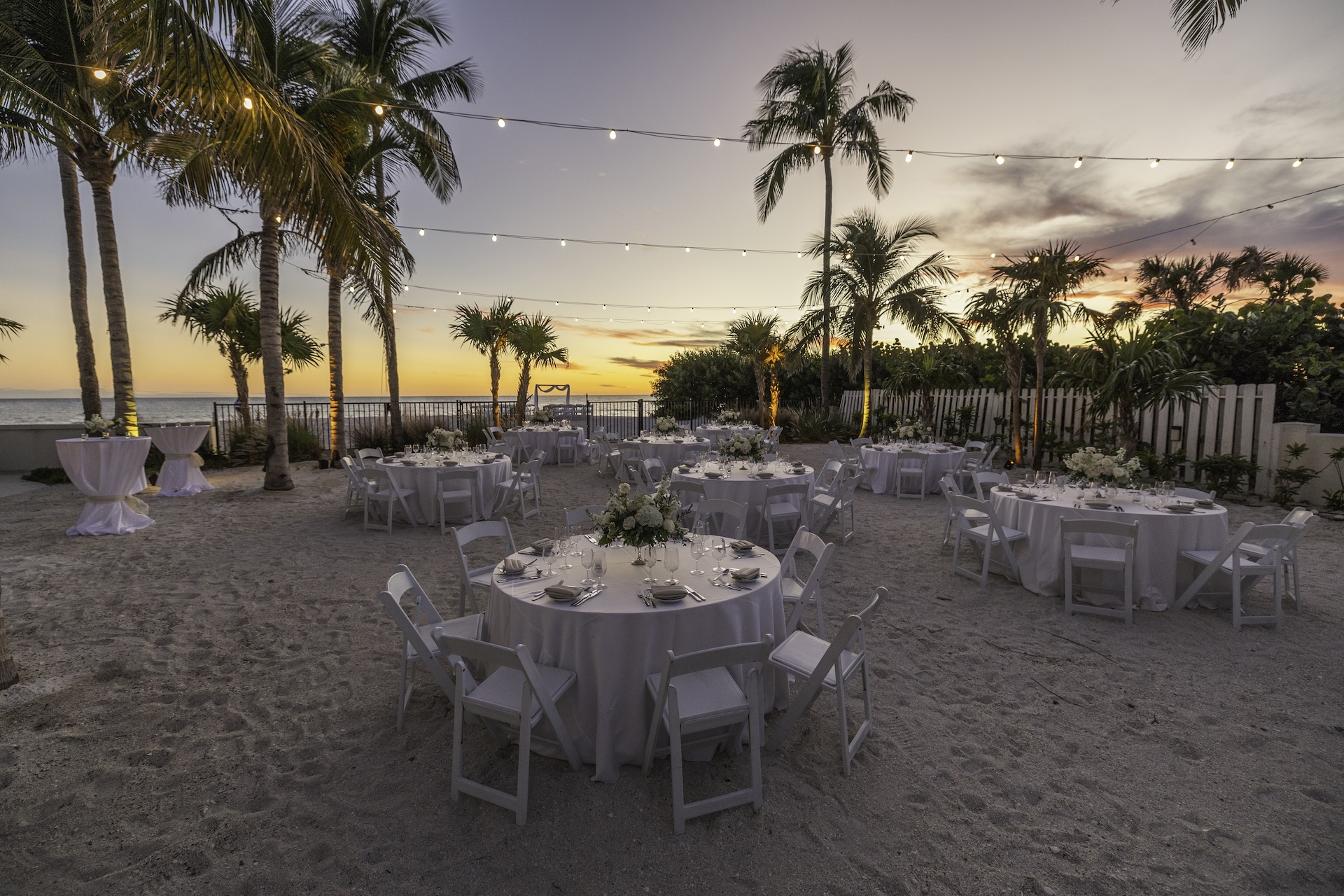 Outdoor beachside event setup at sunset with round tables and white chairs on sand, string lights overhead, palm trees, floral centerpieces, and the ocean visible in the background.