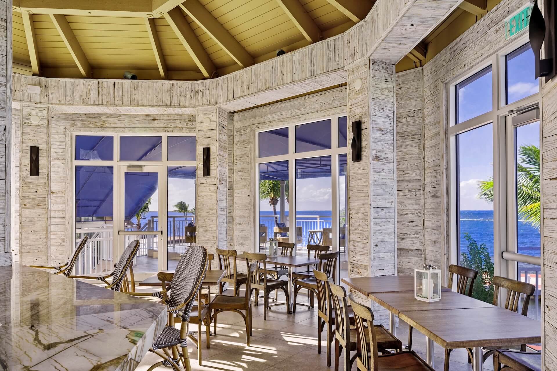 Bright, airy restaurant with wooden tables and chairs, large windows and doors showcasing a view of the ocean, palm trees, and blue sky, with natural light streaming in.
