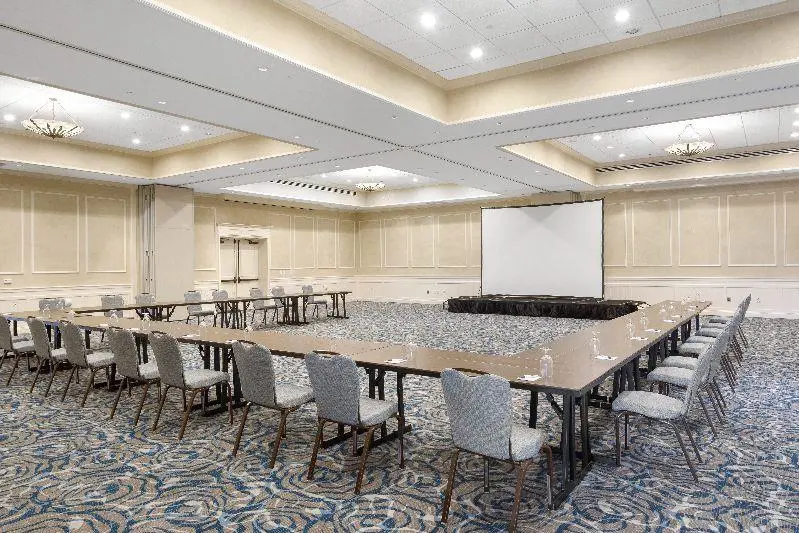 A large, well-lit conference room with U-shaped tables and gray chairs, facing a projector screen. The room has patterned carpet, beige walls, and ceiling lights. Water glasses are placed on the tables.