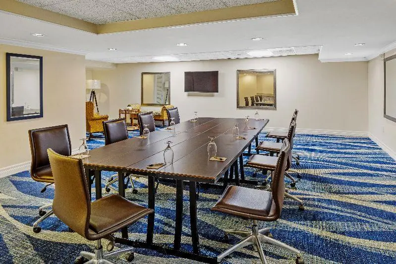 Modern conference room with a rectangular table, eight brown chairs on wheels, water glasses, notepads, a wall-mounted TV, blue patterned carpet, beige walls, and large mirrors.