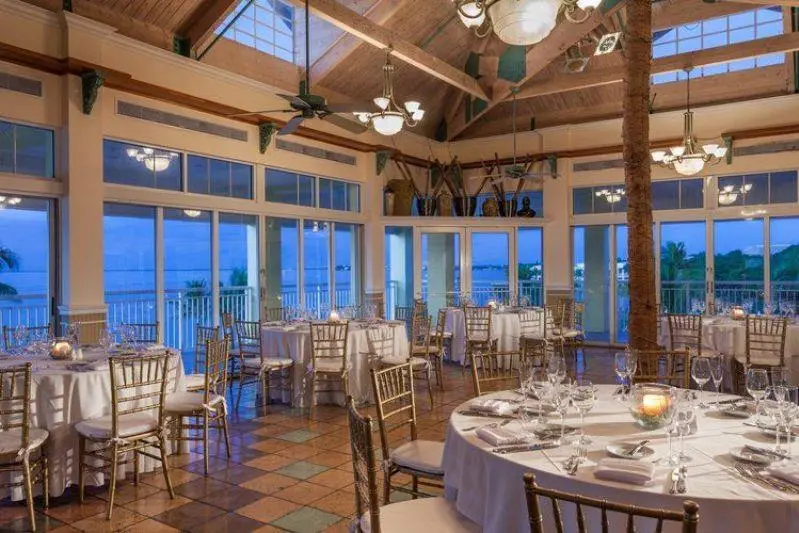 Elegant dining room with round tables set for an event, gold chairs, large windows offering a view of water and palm trees, and high wooden ceilings with fans and hanging lights.