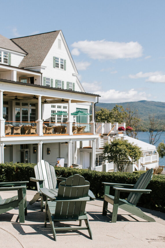 Green Adirondack chairs on a patio overlook a large white house with a spacious deck and outdoor seating. The background features a lake and mountains under a clear blue sky.