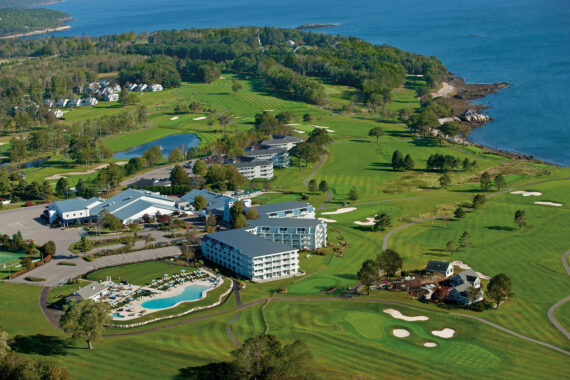 Aerial view of a resort by the ocean, featuring green lawns, a golf course, a swimming pool, and several buildings surrounded by lush trees and rocky shorelines. The sea is visible in the background.
