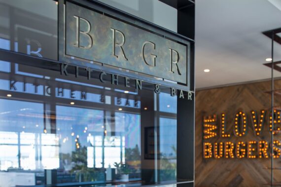 Sign for BRGR Kitchen & Bar is displayed on a window with reflections inside a restaurant. In the background, a lighted sign reads LOVE BURGERS on a wooden wall. Bright, modern interior is partially visible.