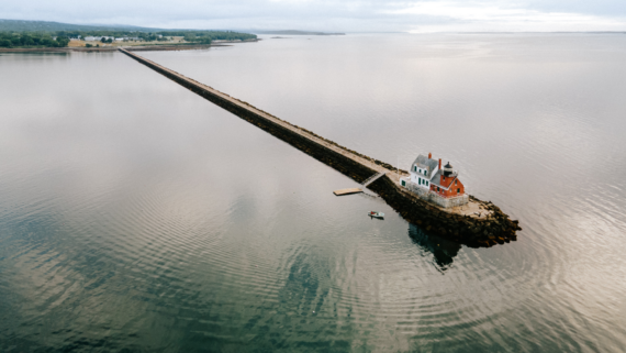 Aerial view of a narrow stone breakwater extending into calm water, leading to a small lighthouse with a red-roofed building. A boat is moored nearby. The surrounding landscape is tranquil and partly cloudy.