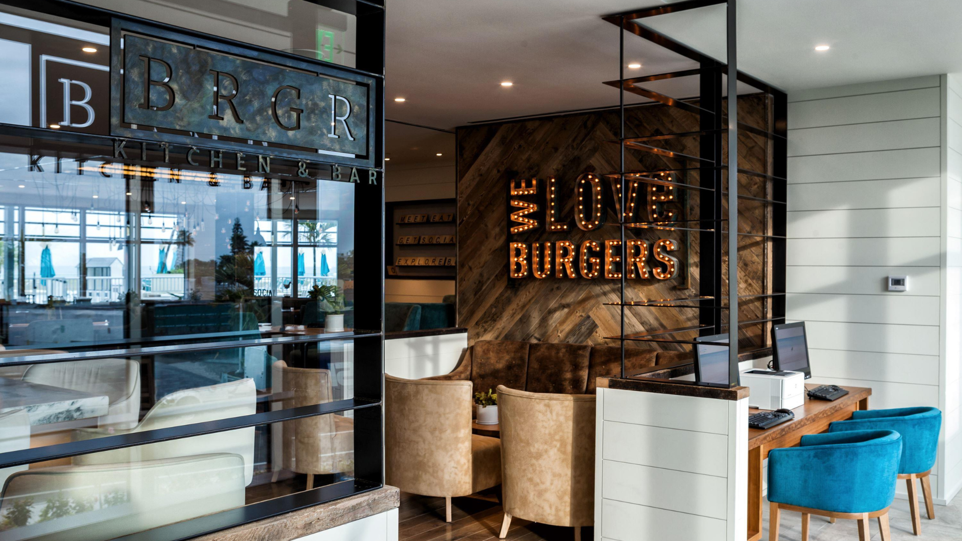 A modern restaurant interior featuring a sign reading WE LOVE BURGERS in lights on a wooden wall. The space includes tables, chairs, and a counter with computers, blending comfort with sleek design. The restaurant is called BRGR Kitchen & Bar.