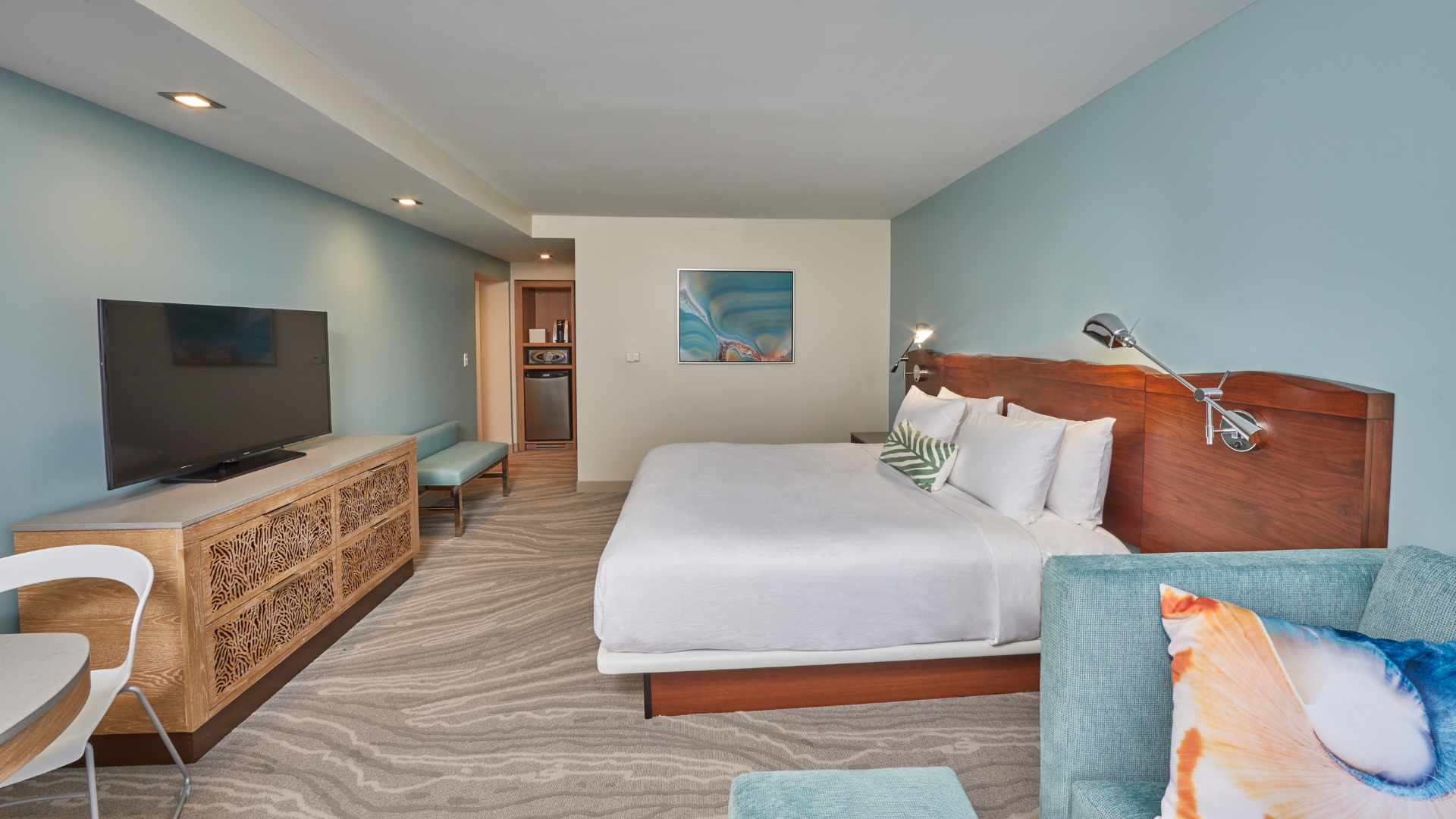 Hotel room with a king-size bed, white linens, and turquoise accents. Theres a TV on a wooden console, a teal armchair, and coastal-themed decor. Walls are light blue, and the carpet has a subtle wave pattern.
