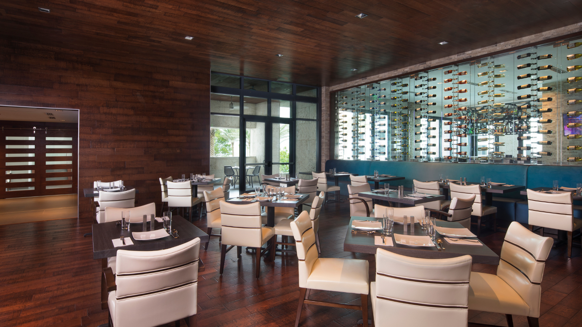 A modern restaurant interior with wooden floors and ceiling, white cushioned chairs, and dark wooden tables set for dining. A glass wine display adorns the back wall. Large windows provide natural light, creating a warm and inviting atmosphere.
