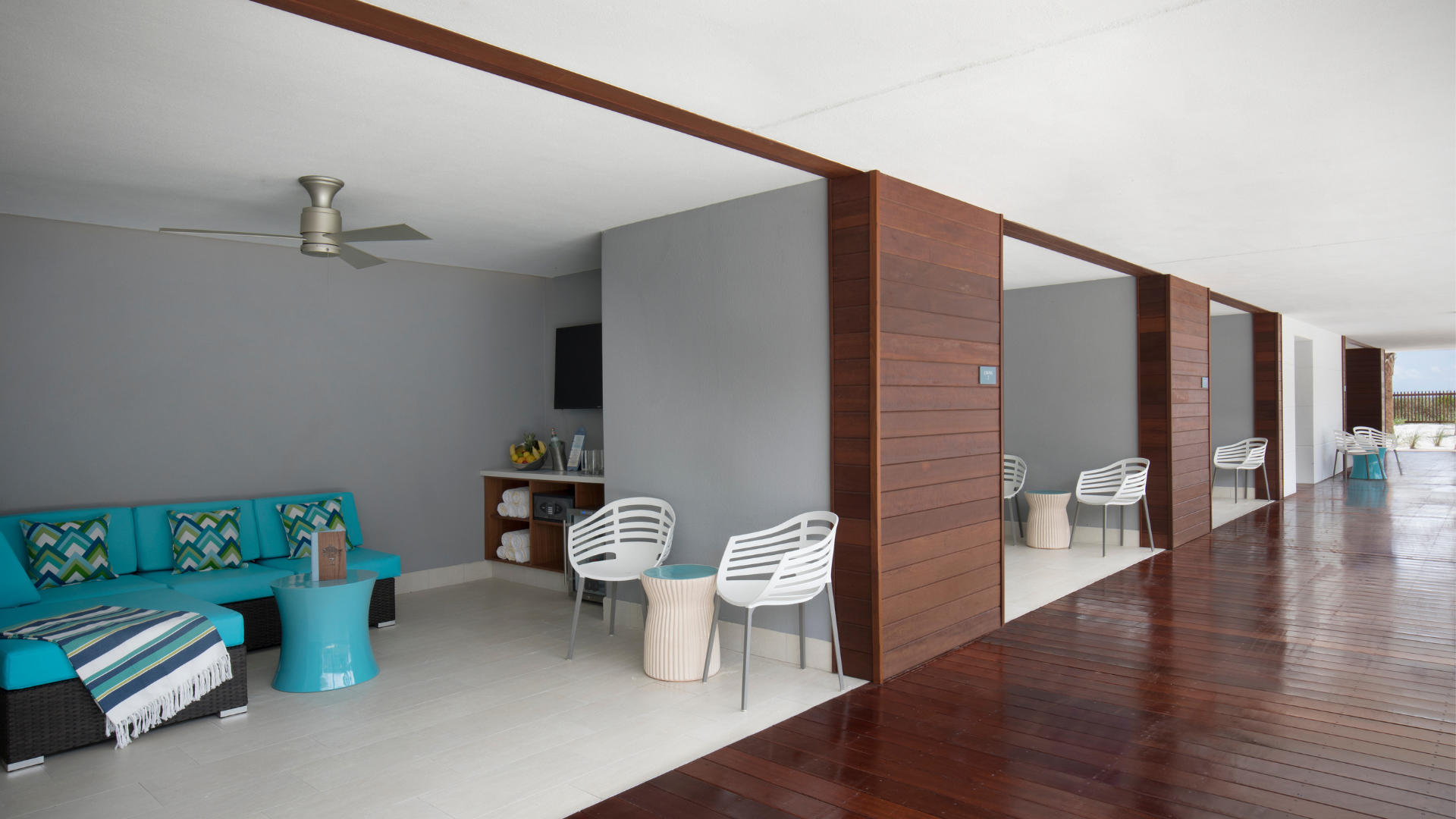 A modern lounge area with turquoise seating and colorful cushions, adjacent to a wooden deck. White chairs and small tables occupy the deck space, separated by wooden partitions. A ceiling fan and neutral walls complete the contemporary look.