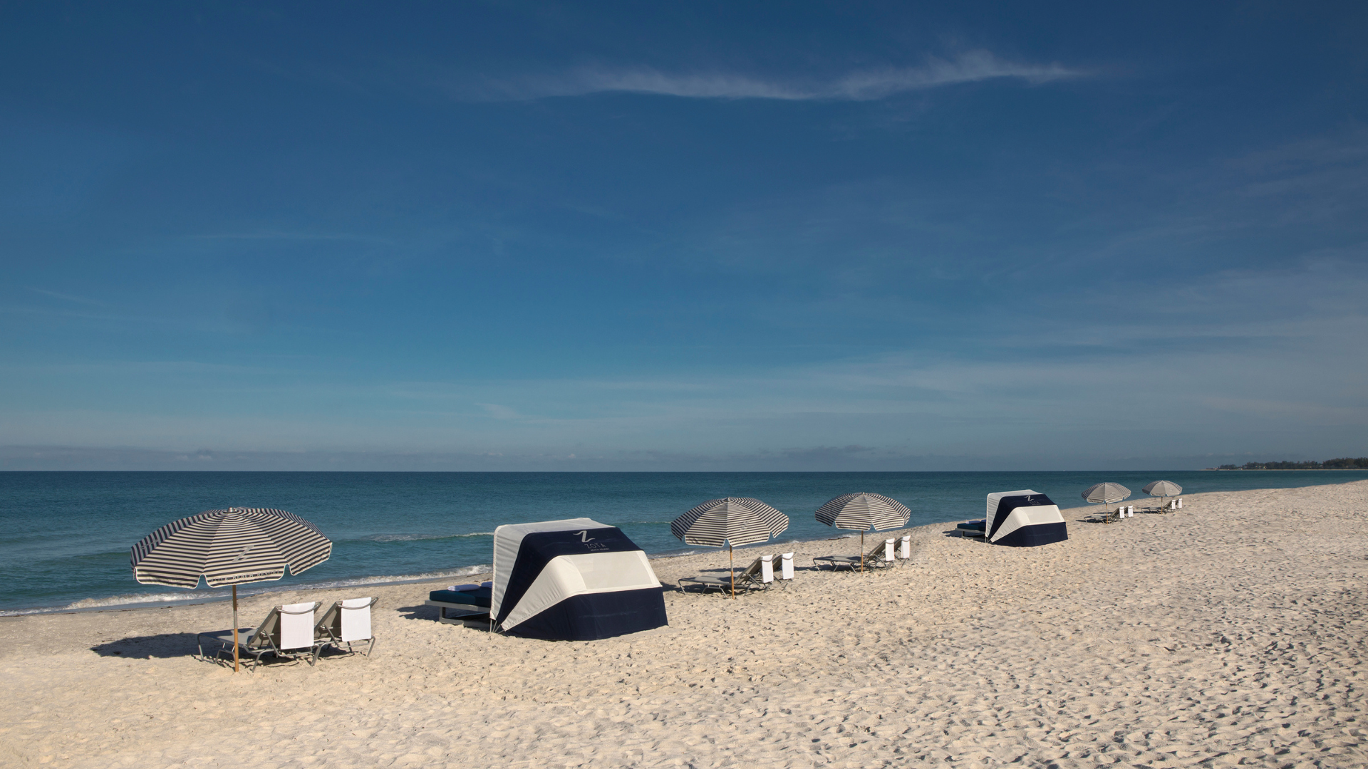 A serene beach scene with lined-up lounge chairs and blue-and-white striped umbrellas facing the ocean. The beach is empty, and the sky is clear with a few scattered clouds.