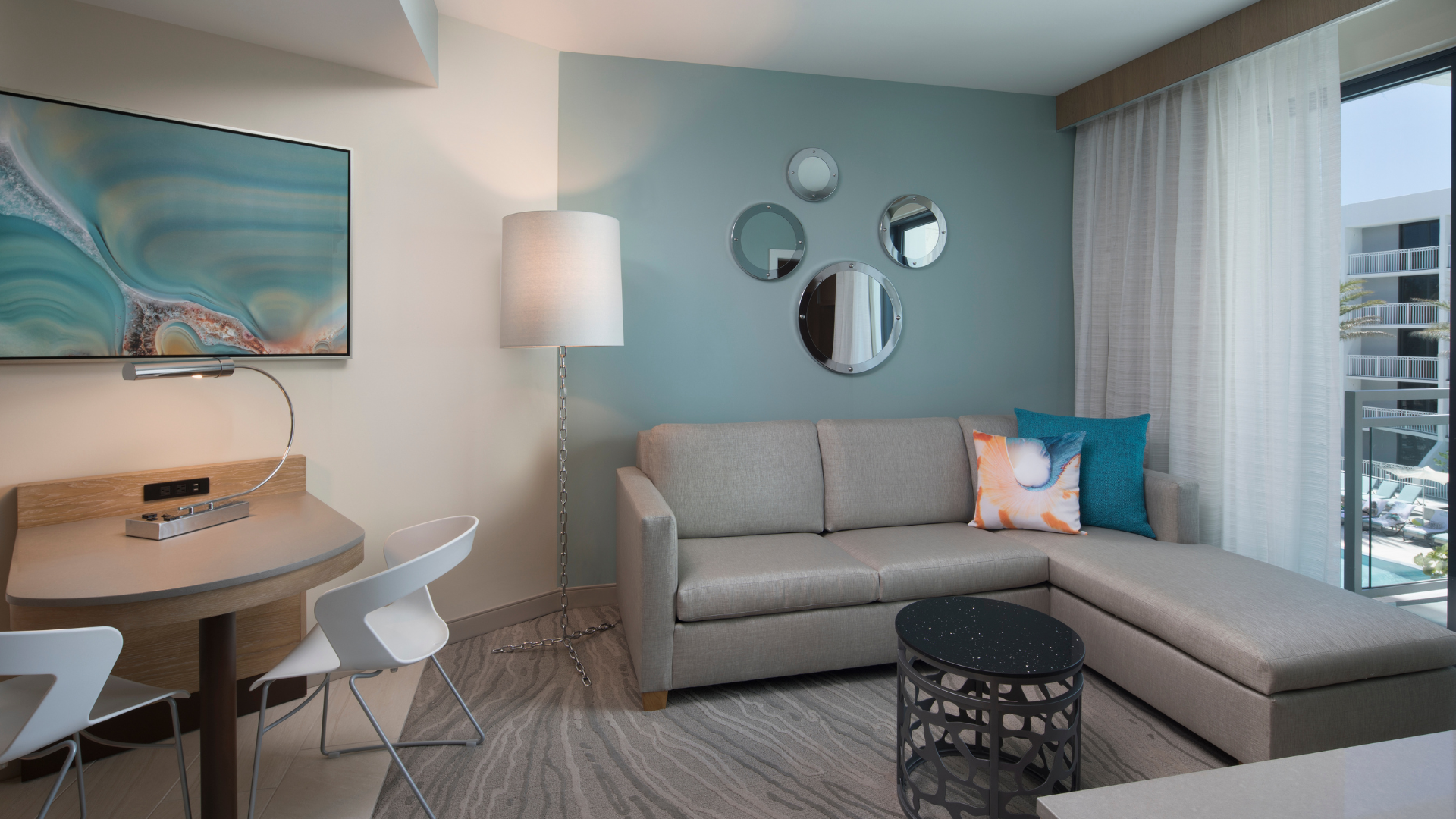Modern hotel room with a gray sectional sofa, teal accent wall with circular mirrors, and a flat-screen TV. A small desk with chairs and a lamp is on the left. Large windows with sheer curtains allow natural light to fill the room.