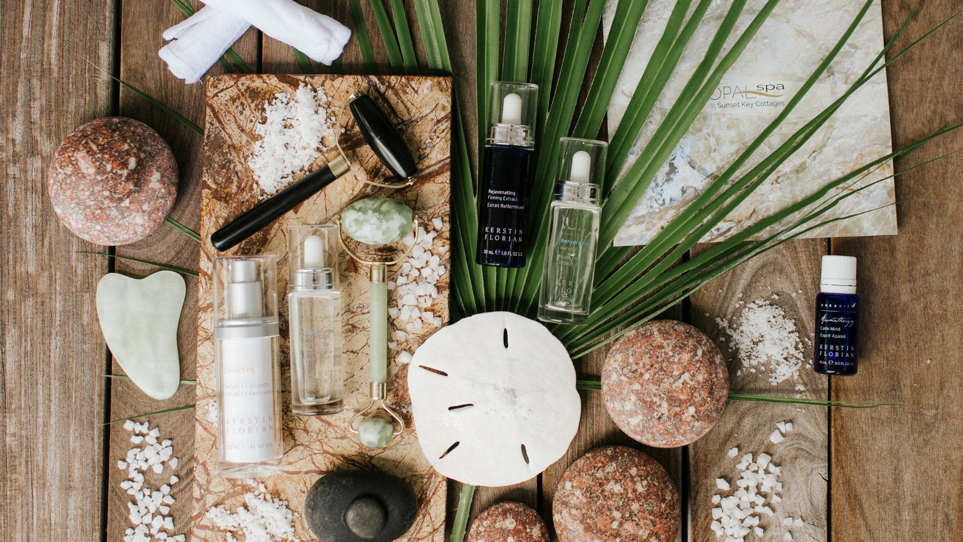 A collection of skincare products and stones on a natural surface, accompanied by a sand dollar, a palm leaf, and small white pebbles. Various spray and serum bottles are arranged artfully, creating a serene spa-like setting.