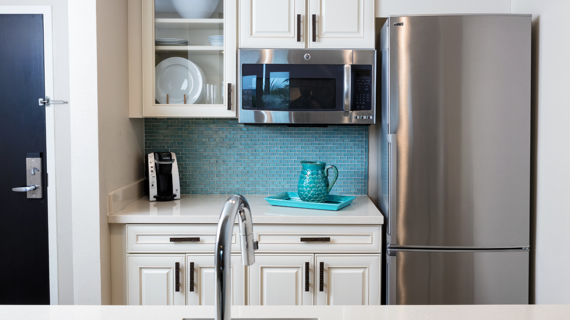 A modern kitchen with a stainless steel refrigerator, microwave, white cabinets, and a coffee maker. A teal jug sits on a matching tray on the countertop, and theres a blue tiled backsplash. A silver faucet is visible in the foreground.