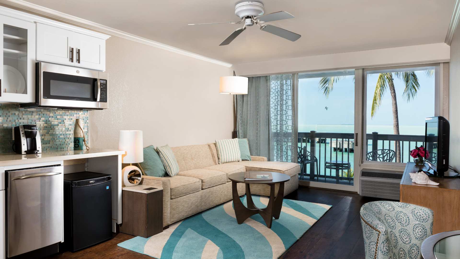 A modern hotel room features a small kitchenette with stainless steel appliances, a beige sofa, and a round coffee table on a turquoise rug. A sliding glass door opens to a balcony with a view of palm trees and the ocean.